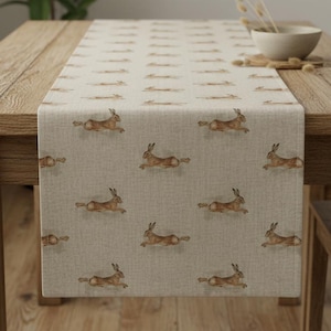 Könnte beinhalten: Beiger Tischläufer mit einem sich wiederholenden Muster aus braunen Hasen in Laufposition. Der Läufer wird auf einem Holztisch präsentiert, der sein Design und seine Textur zeigt. Die Gesamtästhetik ist rustikal und von der Natur inspiriert.