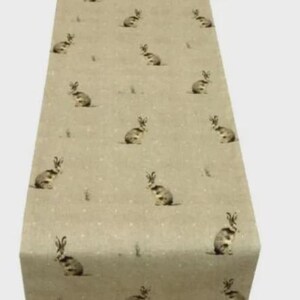 Può includere: Runner da tavolo beige con un motivo ripetuto di conigli in stile acquerello e piccoli puntini bianchi. I conigli sono in varie pose, aggiungendo un tocco stravagante. Il runner è realizzato in un materiale strutturato.