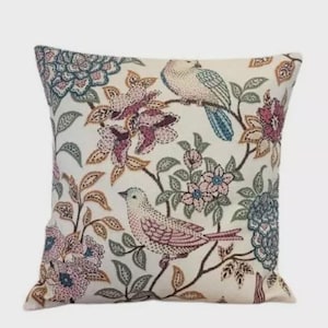 Peut inclure: Coussin carré avec un fond crème orné d'un motif floral et d'oiseaux. Les oiseaux sont dans des tons de rose, bleu et blanc, perchés sur des branches avec des feuilles et des fleurs roses, bleues et marron.