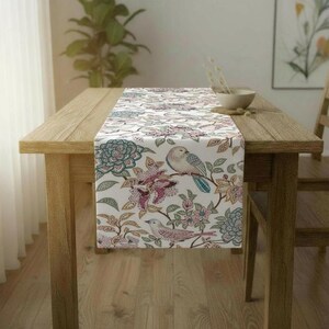 May include: A white table runner with a floral and bird pattern. The design features teal, pink, and brown flowers and birds on branches. The runner is placed on a wooden table.