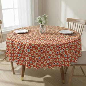 Könnte beinhalten: Eine runde orangefarbene Tischdecke mit einem geometrischen Muster aus bunten Sechsecken bedeckt einen Holztisch. Zwei weiße Teller und eine Vase mit weißen Blumen stehen auf dem Tisch. Zwei Holzstühle umgeben den Tisch.