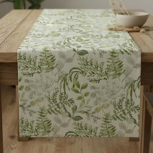 May include: A table runner featuring a botanical leaf design in green and grey hues. The runner is laid on a wooden table. The table has a natural wood finish. A bowl and decorative items are on the table.