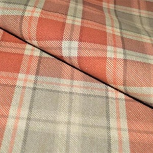 Könnte beinhalten: Nahaufnahme eines karierten Stoffes mit diagonaler Falte. Das Muster zeigt ein Gitter aus sich kreuzenden Linien in Orange-, Grau- und Weißtönen. Der Stoff scheint weich und strukturiert zu sein, geeignet für Kleidung oder Heimdekoration.
