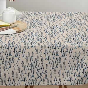 Op de afbeelding: Een beige tafelkleed met een herhalend patroon van blauwe vissen. Het tafelkleed ligt op een tafel met een houten onderstel.