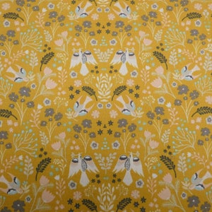 May include: Mustard yellow fabric featuring a repeating pattern of white birds, grey and pink flowers, and green foliage. The design is symmetrical, creating a visually balanced and decorative textile.