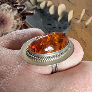 May include: A silver ring featuring a large, oval amber stone. The amber is a warm, translucent orange color with visible inclusions. The ring has a decorative rope-like border around the amber and a simple band.