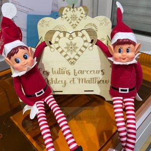 May include: Two red and white striped elf dolls with black boots are sitting on a wooden shelf with a heart design and the text "Les lutins farceurs Ashley et Matthew".
