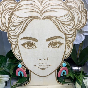 May include: A pair of colourful, geometric earrings with a rainbow arch design. The earrings are made of pink, blue and white clay and are hanging from a wooden mannequin head.