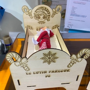 Puede incluir: Una cama de madera para elfos con un diseño de bastón de caramelo y un copo de nieve. La cama tiene un letrero que dice "Le Lutin Farceur De".