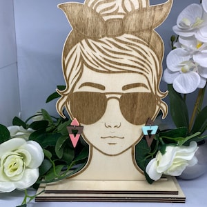 Könnte beinhalten: Ein hölzerner Ohrringständer mit einem Frauenkopf, Sonnenbrille und einem Paar Dreiecksohrringen in Rosa, Blau und Braun.