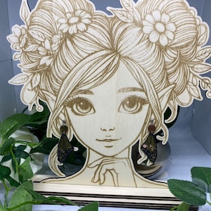 May include: A wooden cutout of a woman with floral hair, wearing butterfly earrings. The cutout is on a wooden stand.