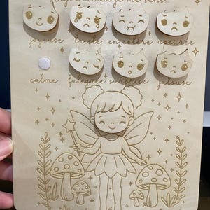 May include: Wooden board with a fairy illustration and a set of interchangeable wooden emotion faces. The board has the text "Emma Aujourd'hui je me sens..." and lists emotions like "joyeuse", "triste", "en colère", "apeurée", "calme", "fatiguée", "surprise", and "jalouse".