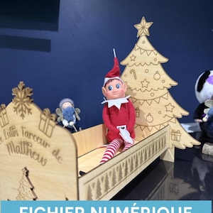 Op de afbeelding: Een houten bed- en kerstboomset met een elfenpop. Het bed heeft gegraveerde details en tekst. De elf draagt een rood pak en gestreepte benen. De kerstboom is versierd met sterren en ornamenten. De tekst "FICHIER NUMÉRIQUE FORMATS SVG & XCS" staat onderaan.