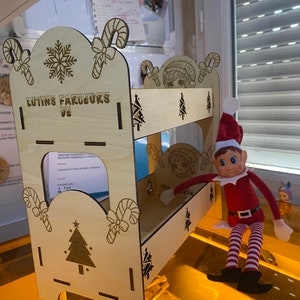 Peut inclure: Une étagère en bois sur le thème de Noël. L'étagère a deux niveaux et est décorée de cannes en sucre, de flocons de neige et de sapins de Noël. L'étagère est étiquetée "Lutins Farceurs De". Une poupée d'elfe de Noël est assise sur l'étagère supérieure.