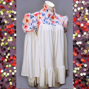 May include: A cream-colored dress with a high neck and floral print on the shoulders and neckline. The dress features a ruffled hem and long, flared sleeves. The floral pattern includes red, pink, and blue flowers.