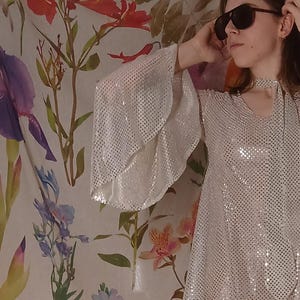 May include: A silver sequinned top featuring wide bell sleeves and a high neckline with a tie detail. The fabric is covered in small, reflective metallic discs that create a shimmering effect across the entire surface of the garment.