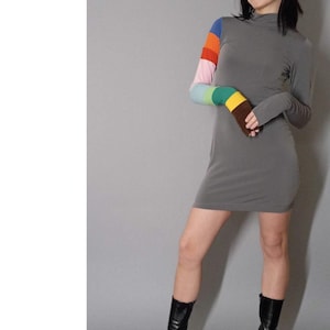 May include: A gray mini dress with a high neck and long sleeves. The sleeves have a colorful striped pattern. The model is wearing black ankle boots.