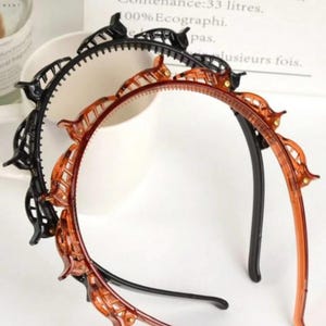 May include: Two hair headbands, one black and one amber, with a series of decorative clips. The headbands are made of plastic and are designed to hold hair in place. The background is white.
