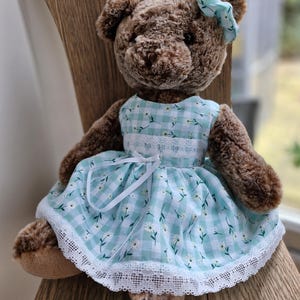 May include: A brown teddy bear wearing a light blue gingham dress with white daisy accents and a matching bow. The dress has a white lace trim. The bear is sitting on a wooden surface.