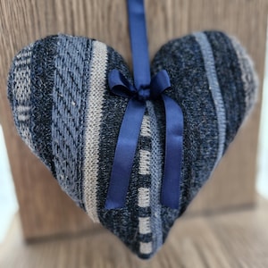 May include: A heart-shaped decoration crafted from knitted fabric in various shades of blue, grey, and beige. A navy blue satin ribbon is tied in a bow at the top, with a loop for hanging. The knitted pattern features stripes and textured areas.