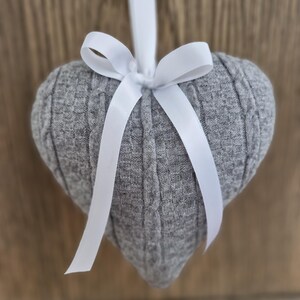 May include: A grey, heart-shaped decoration made from knitted fabric, with a white ribbon bow for hanging. The ornament features a cable knit texture and is set against a blurred wooden backdrop.