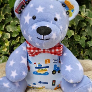 May include: A plush teddy bear made of light blue fabric with a white star pattern. The bear features a red and white gingham bow tie and a torso printed with various vehicles, including a boat, tractor, and traffic light. It has black eyes.