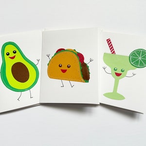 May include: Three greeting cards with illustrations of a smiling avocado, a smiling taco, and a smiling margarita with a lime wedge. The cards are white with green, brown, yellow, red, and blue accents.