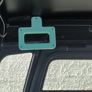 Puede incluir: Un soporte rectangular de color verde azulado claro, con una abertura central y orificios de montaje, está montado en el techo interior de un vehículo. El soporte está situado cerca del parabrisas y parece ser de plástico. El interior del vehículo es negro.