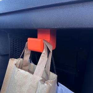 Può includere: Uncino di plastica arancione appeso alla parte posteriore di un sedile auto, che tiene due sacchetti di carta marroni e un sacchetto di carta bianco.