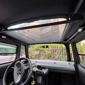 Può includere: Vista interna di una piccola auto nera con tetto trasparente. Sono visibili il volante, il cruscotto e i finestrini laterali. L'auto è parcheggiata all'esterno, con vista su edifici e alberi.