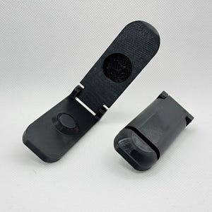 Può includere: Due supporti per telefono neri stampati in 3D. Un supporto è aperto, mostrando un'indentazione circolare e una cerniera. L'altro supporto è chiuso. Entrambi i supporti hanno una superficie strutturata e bordi arrotondati. I supporti sono su una superficie bianca.