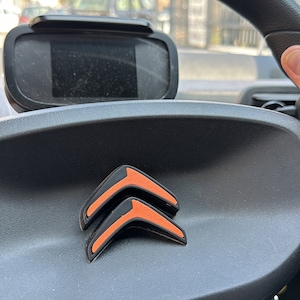 Op de afbeelding: Close-up van een zwart lederen stuurwiel met een zwart en oranje Citroën-logo. Het stuurwiel bevindt zich in een auto.