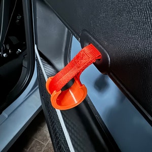 Può includere: Un gancio per portiera di auto stampato in 3D di colore arancione brillante con un cinturino nero attaccato a un pannello di portiera di auto nero.