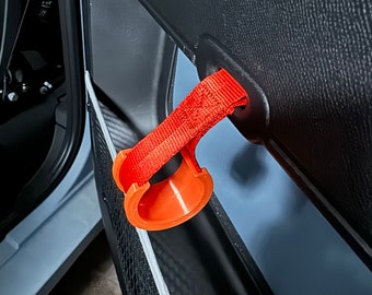 Może przedstawiać: Pomarańczowy, trójwymiarowy haczyk do drzwi samochodowych, wydrukowany w technologii 3D, z czarnym paskiem przymocowanym do czarnego panelu drzwi samochodowych.