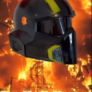 Op de afbeelding: Een zwarte helm met een gele streep en een zwart vizier. De helm staat voor een vurige achtergrond met de tekst "Be Helldivers" in gele letters.