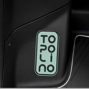 Puede incluir: Un emblema rectangular verde claro y negro con la palabra "TOPOLINO" en texto blanco estilizado. El emblema está sobre un fondo negro, con un borde blanco.
