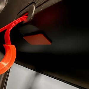 Op de afbeelding: Een close-up van een auto-interieur met een rode handgreep en een bijpassend rood, ruitvormig accentstuk. De handgreep is bevestigd aan een zwart oppervlak en het accentstuk is op hetzelfde oppervlak bevestigd.