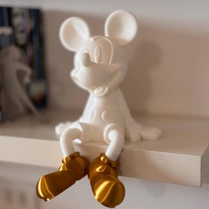 Peut inclure: Figurine blanche de Mickey Mouse assis sur une étagère avec des chaussures dorées.