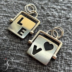 May include: Two silver-toned pendants with a rotating square design. One pendant spells "LE" with a gold-colored background, the other has a "V" and a heart. Each has a small loop for a chain.