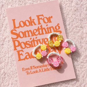 May include: A pink notebook with the text "Look For Something Positive Each Day" and the phrase "Even If Somedays You Have To Look A Little Harder." Several white beaded rings with yellow and pink smiley face charms are arranged on the notebook.