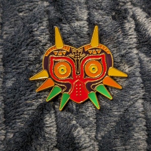 May include: A gold, red, green, and purple enamel pin of the Majora's Mask from the Legend of Zelda video game series.