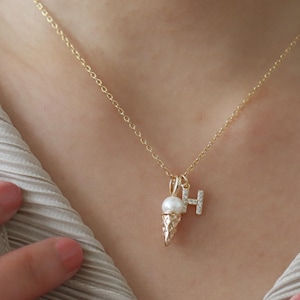 May include: A gold-colored necklace with a pendant shaped like an ice cream cone and the letter "H". The ice cream cone pendant has a pearl and is adorned with small, clear crystals. The necklace is worn on a person's neck.