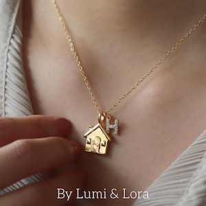 May include: A gold necklace with a house pendant and a letter "H" charm. The house pendant has a door and windows. The necklace is worn on a person's neck. The text "By Lumi & Lora" is at the bottom of the image.