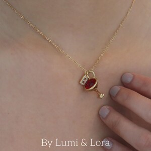 Könnte beinhalten: Goldfarbene Halskette mit einem Anhänger in Form eines Cocktailglases mit einem roten Edelstein und kleinen, klaren Edelsteinen. Die Halskette wird um den Hals einer Person getragen. Der Text "By Lumi & Lora" ist unten im Bild zu sehen.