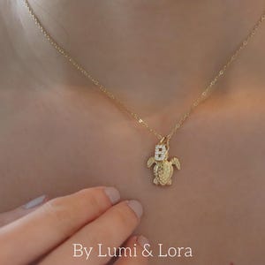 May include: A gold necklace featuring a turtle pendant with a letter "B" and small, clear stones. The necklace is worn. The text "By Lumi & Lora" is at the bottom of the image. The necklace is a delicate piece of jewellery.