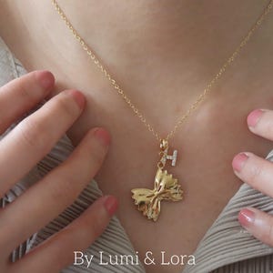 Pode incluir: Um colar dourado com um pingente de laço e um pequeno berloque com a letra "H". O colar é usado ao redor do pescoço. A imagem também inclui o texto "By Lumi & Lora".