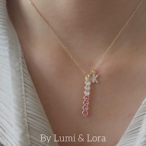 Puede incluir: Collar dorado con un colgante de piedras preciosas en forma de corazón en tonos rosa y blanco, y un charm con la letra "K" engastada con diamantes. El collar se lleva sobre una camisa blanca. El texto "By Lumi & Lora" es visible.