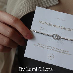 Op de afbeelding: Een witte kaart met de tekst "LUMI & LORA MOEDER EN DOCHTER" en "Deze kleine hartjes zijn onlosmakelijk verbonden, zoals moeder en dochter, voor altijd en eeuwig." De kaart toont ook een zilveren armband met twee met elkaar verbonden hartbedels.