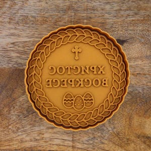 May include: A round, tan-colored cookie cutter with a detailed design. The center features text and three decorated Easter eggs. A cross is at the top, and the design is framed by a braided border.