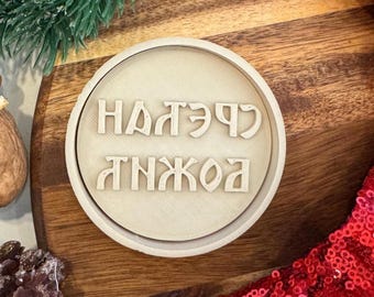 May include: A round, light-colored wooden stamp with the text "НАДЭРЧ АНЖОА" in a stylized font. The stamp is set against a wooden surface, with a sprig of evergreen and red sequined fabric in the background.
